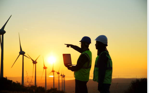 Energy Photo Two workers standing next to each other, one is holding a computer and pointing to wind turbines in the background.