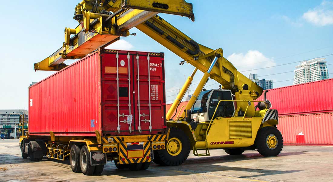 A construction vehicle loading a shipping freight container on to an 18 wheel truck flat bed.